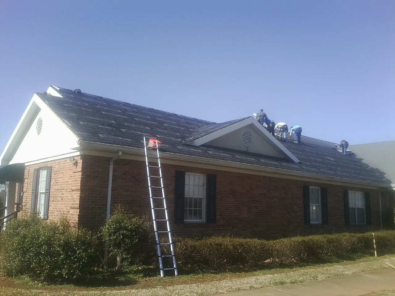 Crew tearing shingles off a brick ranch roof with a ladder set — Metal Roof Repair work by our crew in Holcomb